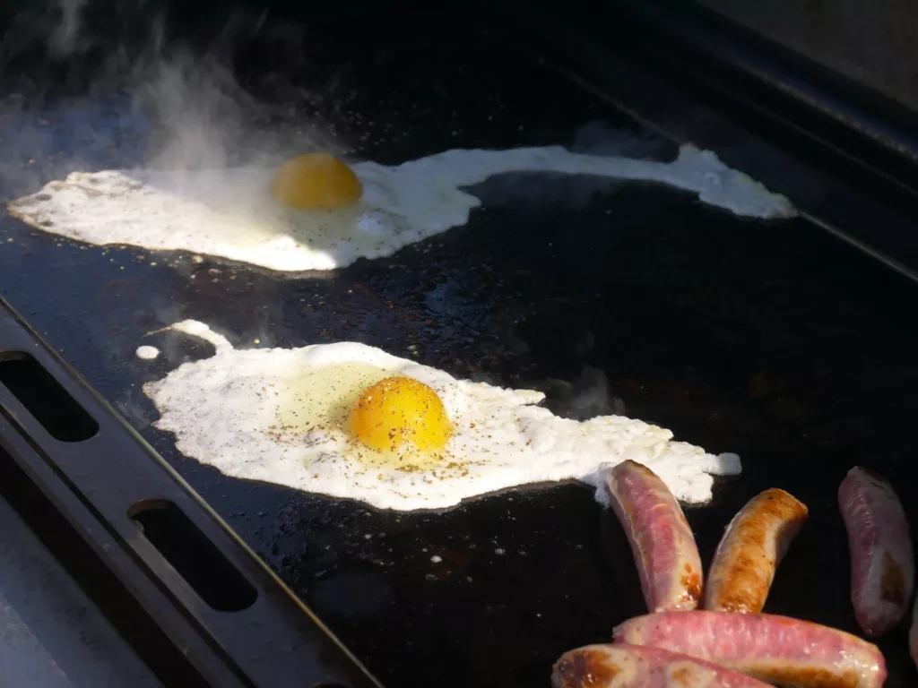 Outdoor brunch cooking on a cast iron griddle with bacon and eggs